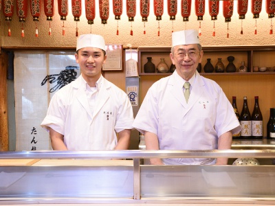 Masahiro Kurisu, the third-generation owner, stands at the counter, and Yuichi, the fourth-generation owner who currently manages the kitchen.
